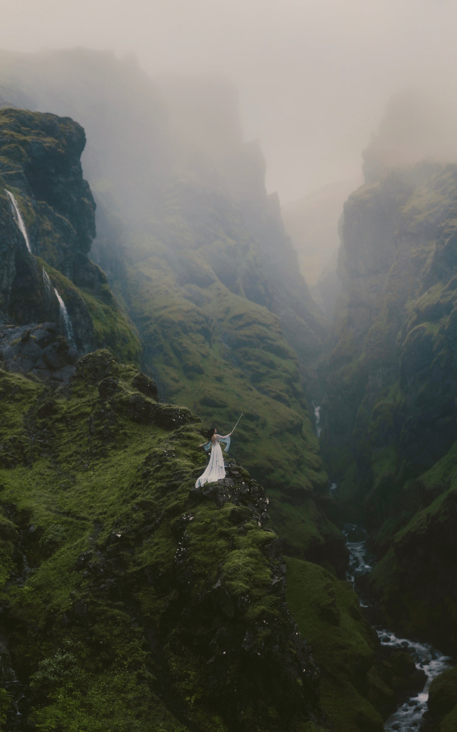 A dramatic landscape photograph of a person in a long dress standing on a mossy green cliffside in a misty mountain canyon with waterfalls and a river below in Iceland.