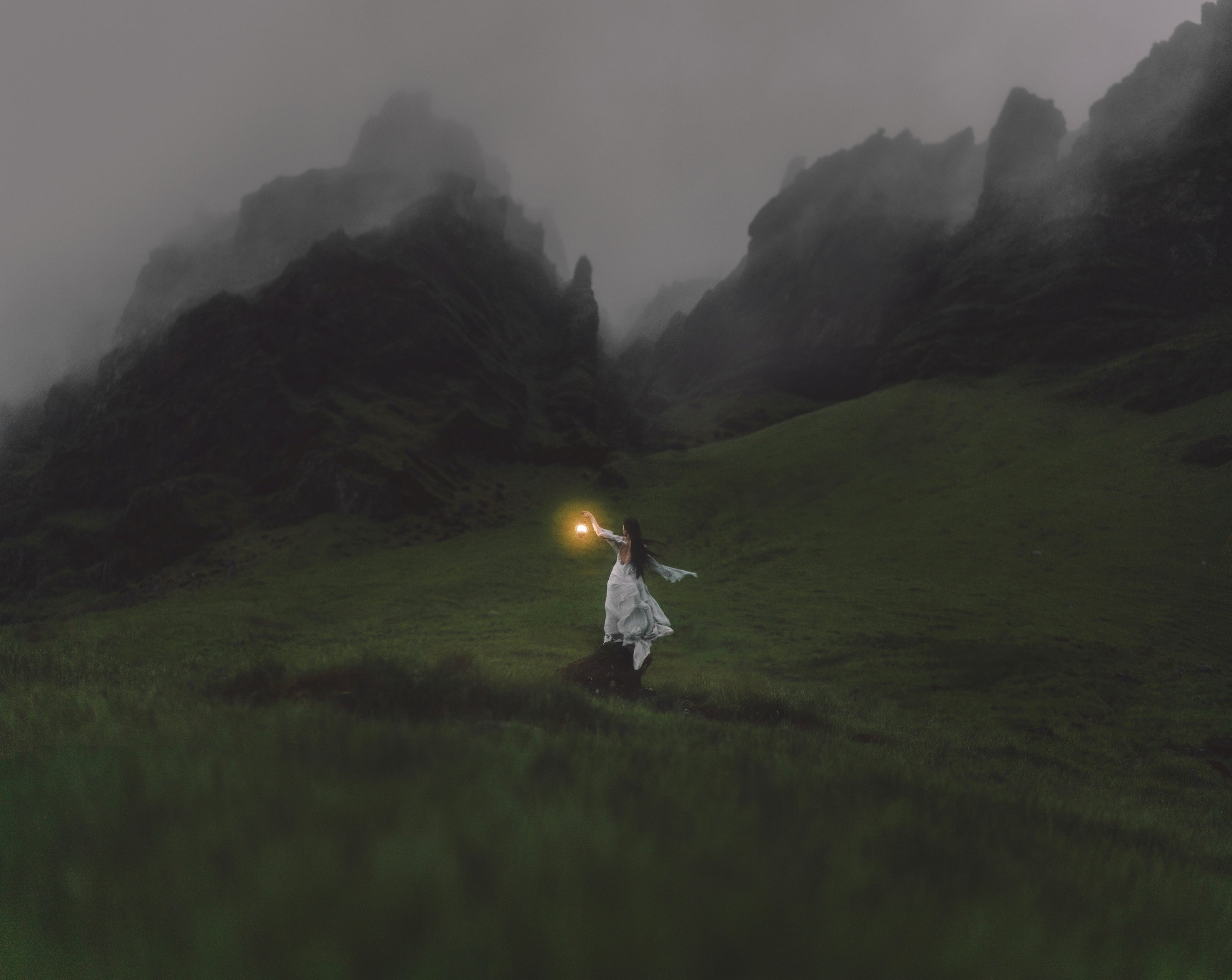 A woman in a long windswept silk dress holds a lantern up to a misty mountain canyon in Iceland