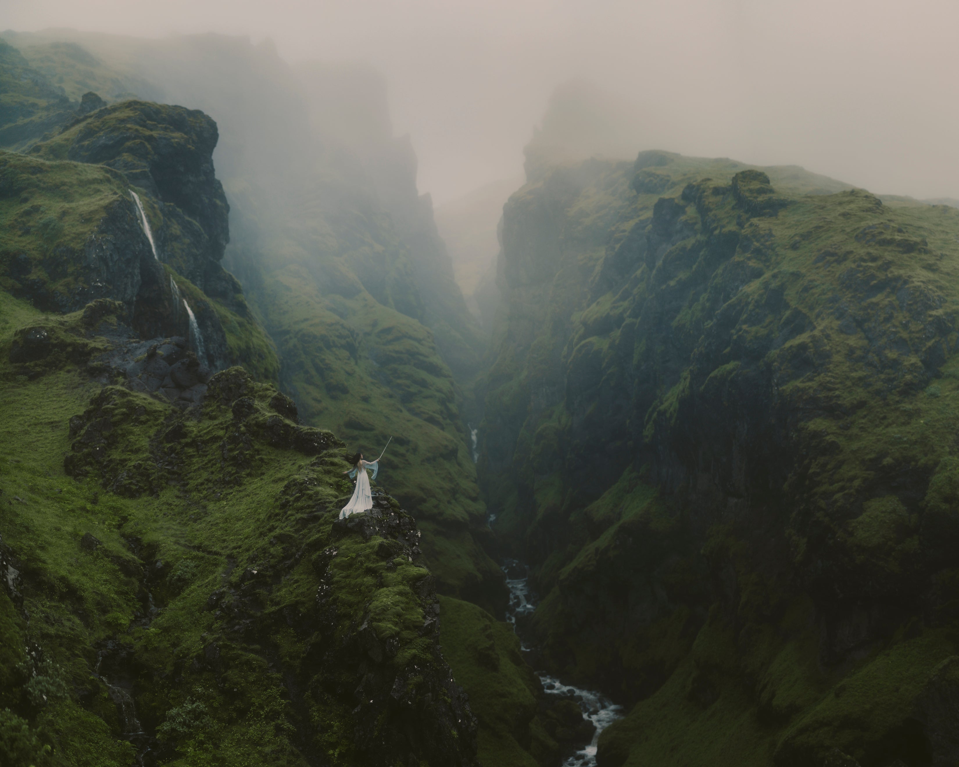 A dramatic landscape photograph in Iceland of a misty green mountain canyon with a waterfall on the left, a winding river in the valley, and a small figure in a white dress standing on a cliff edge.