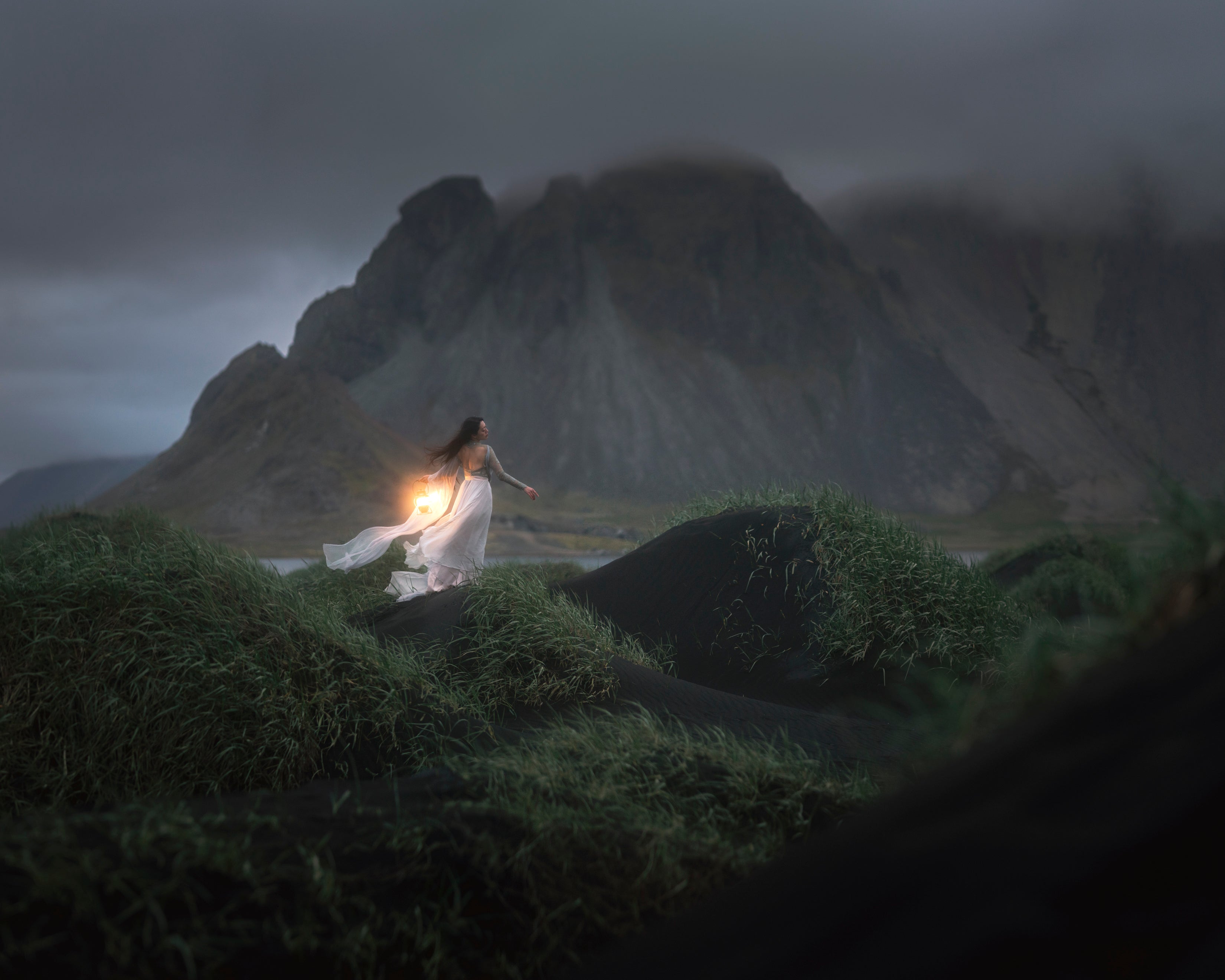 A woman in a flowing white dress stands on a grass dunes in Stokksnes holding a glowing lantern, with dark mountains and a cloudy sky in the background.