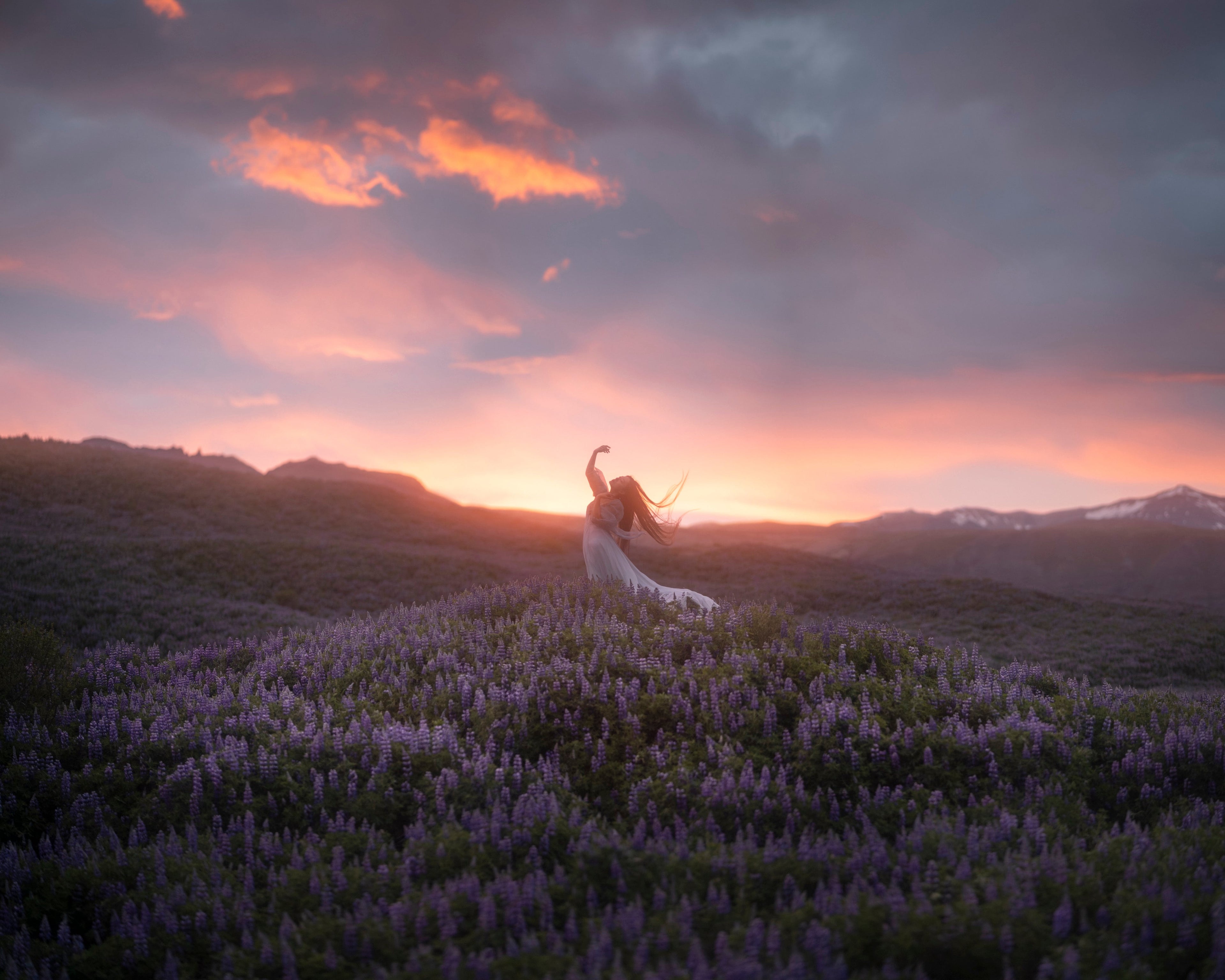 A woman in a flowing dress stands on a hill covered in purple flowers under the midnight sun in Svinafellsjokul, with mountains and a dramatic pink and orange sky behind her.