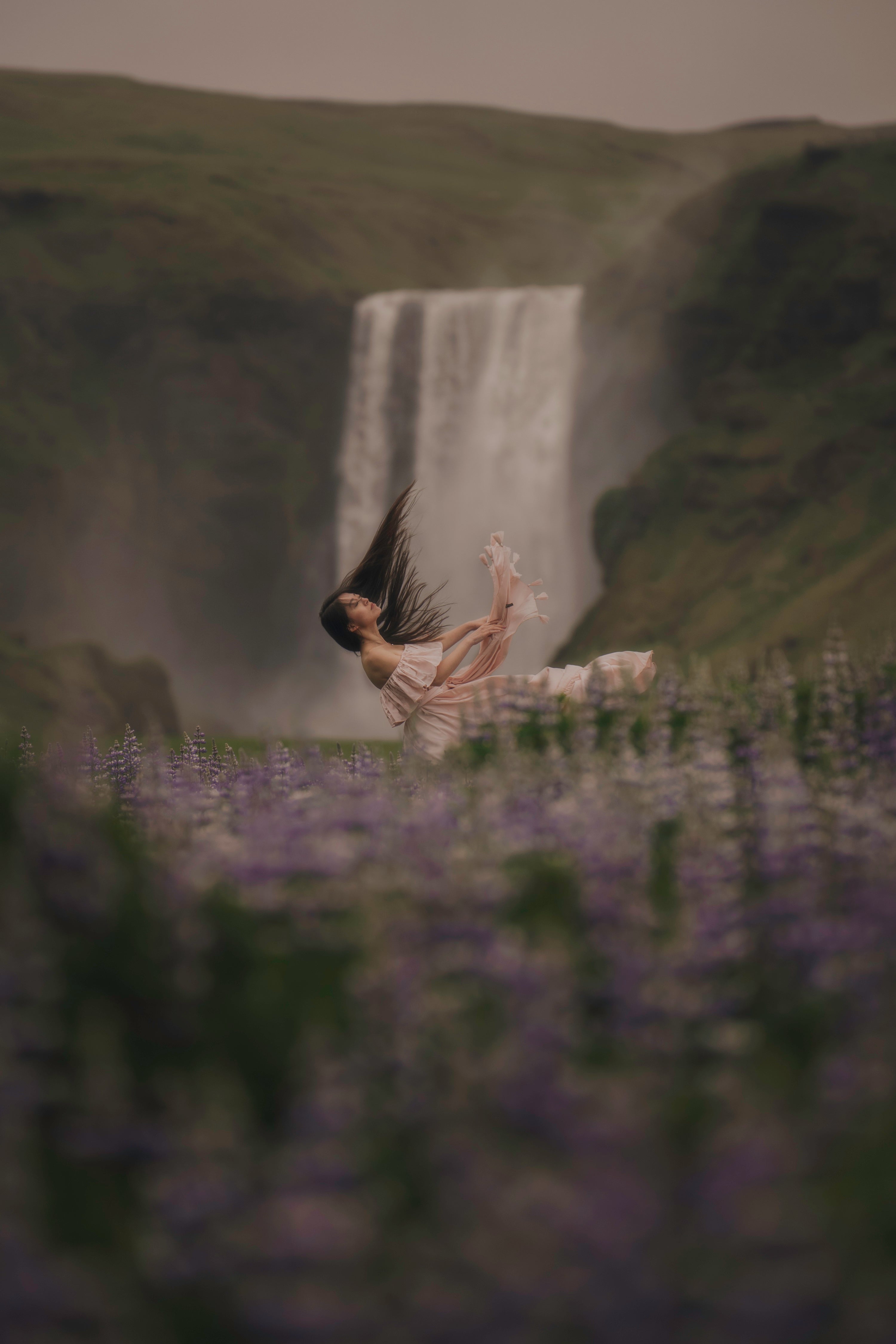 A woman in a flowing dress is posed  falling in front of a tall waterfall named Skogafoss, with blurred purple lupines in the foreground and dark green hills behind her.
