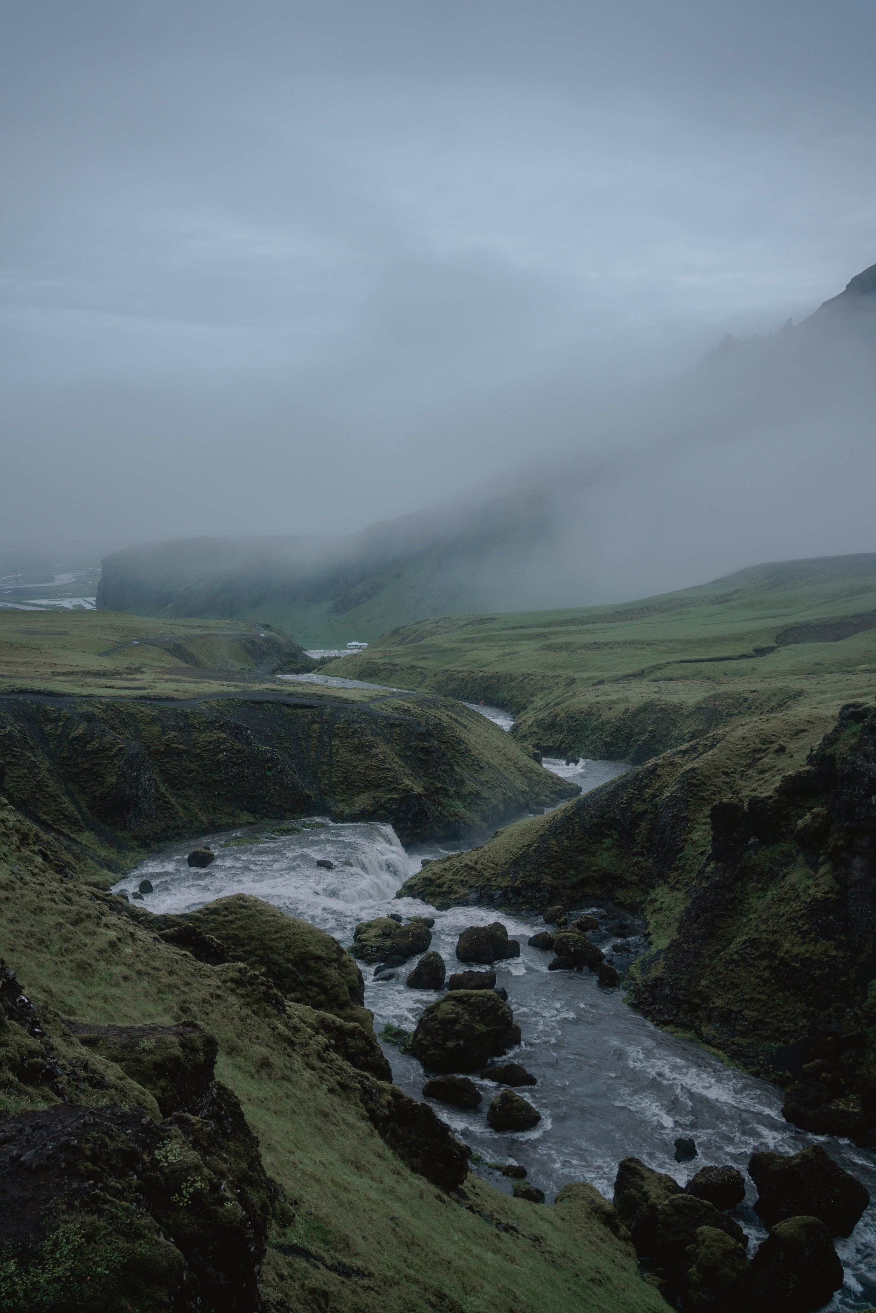 A moody landscape photograph of a winding river flowing through a green, rocky valley above Skogafoss under a heavy, overcast sky with low-lying mist and fog.