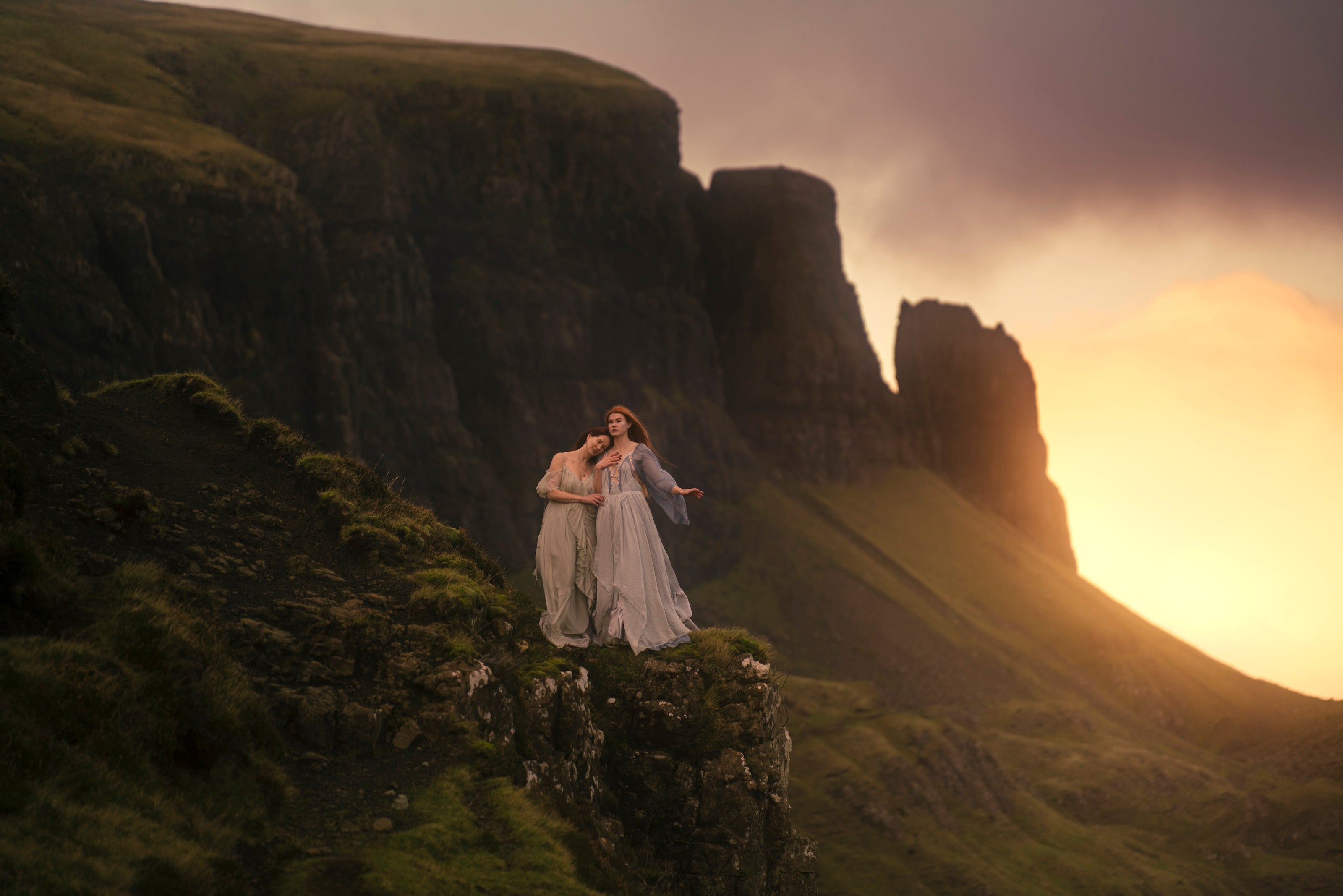 Mother and daughter in flowy dresses standing on a Scottish cliff with a sunset in the background