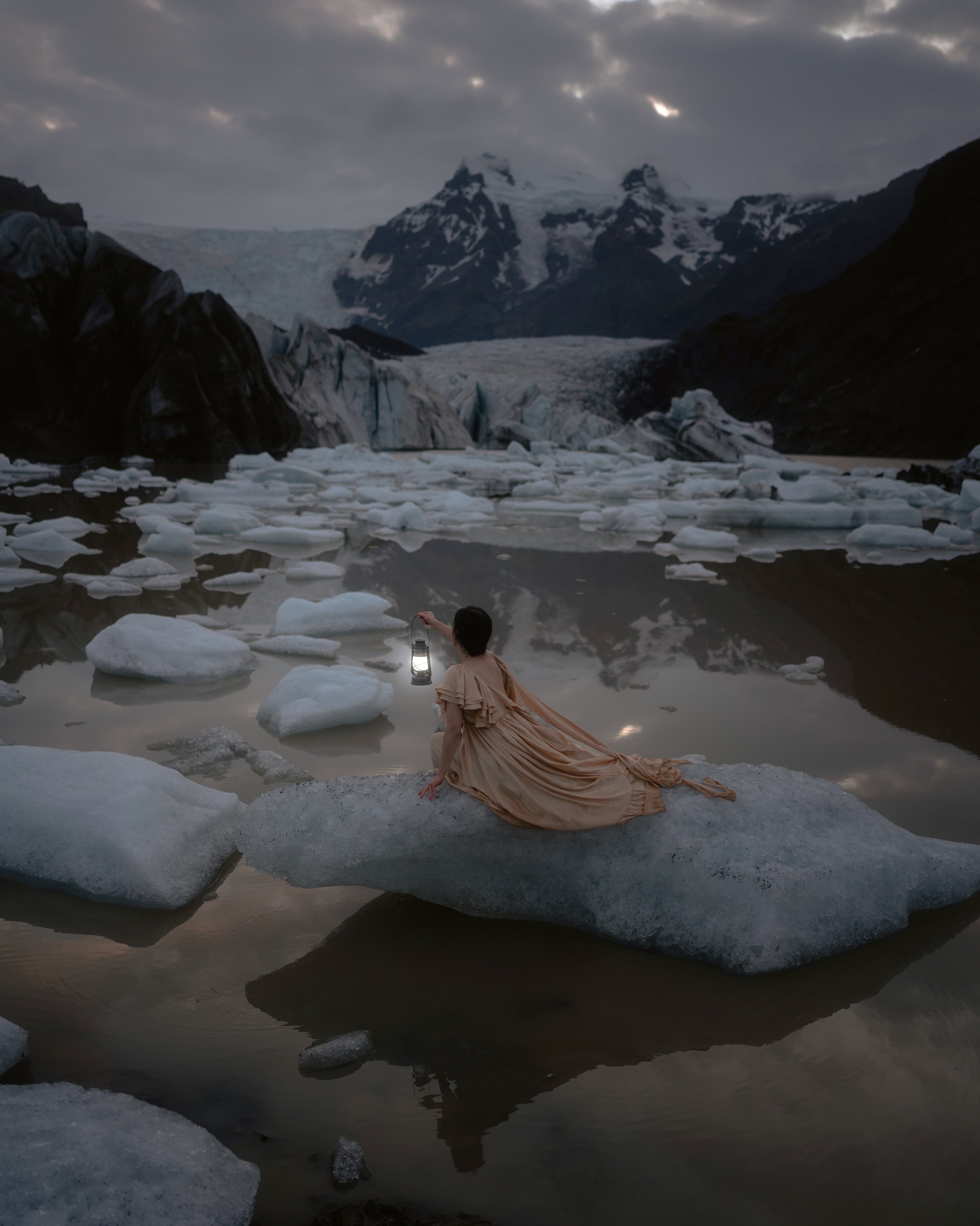 A woman in a flowing dress sits on a large ice floe in Svinafellsjokul glacial lagoon, holding a lantern. Snow-covered mountains and icebergs fill the water under a dark, dramatic sky.