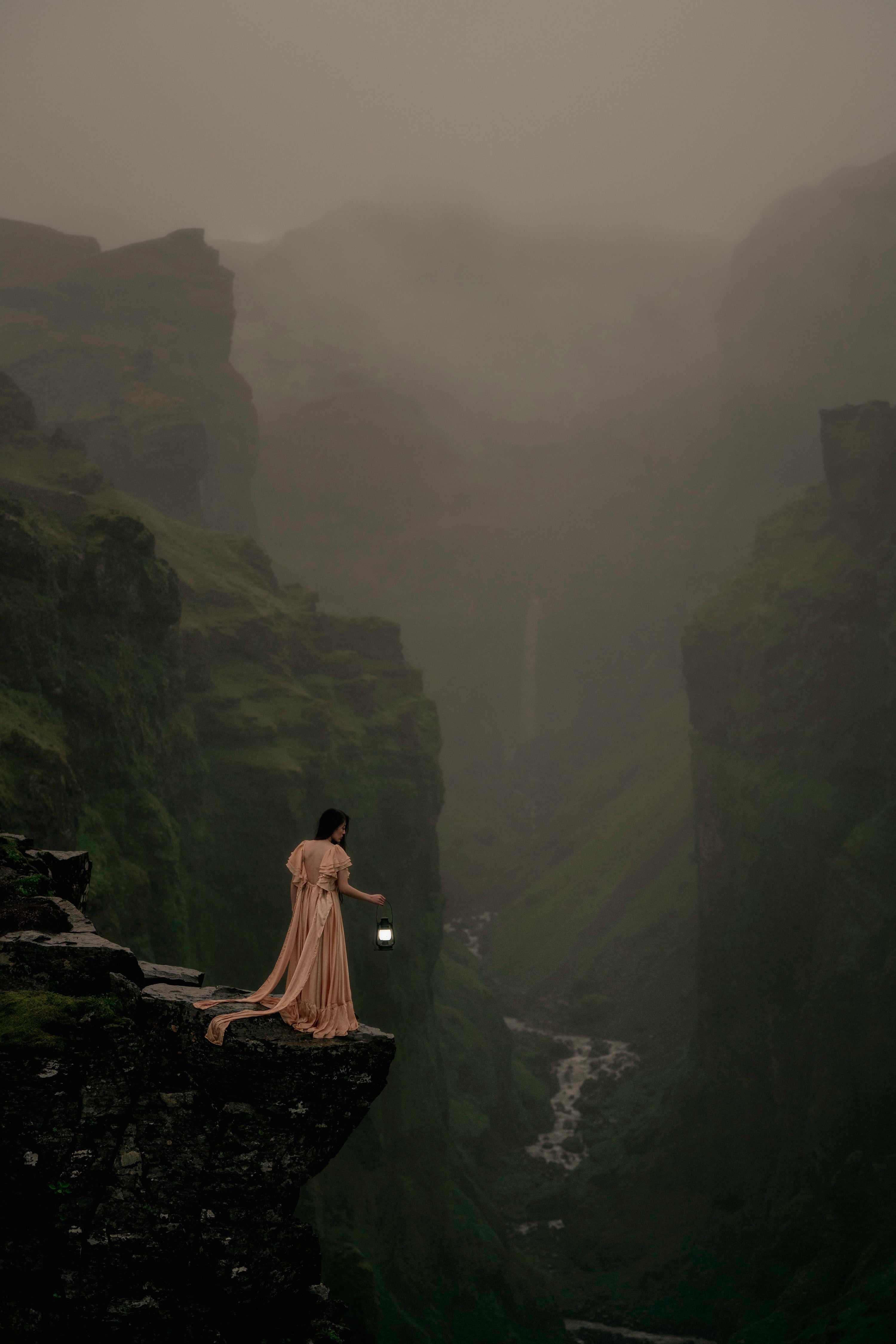 A woman in a long gown stands on a dark cliff edge holding a small glowing lantern, overlooking a misty, dramatic canyon named Mulagljufur with steep rock walls and a waterfall in the distance.
