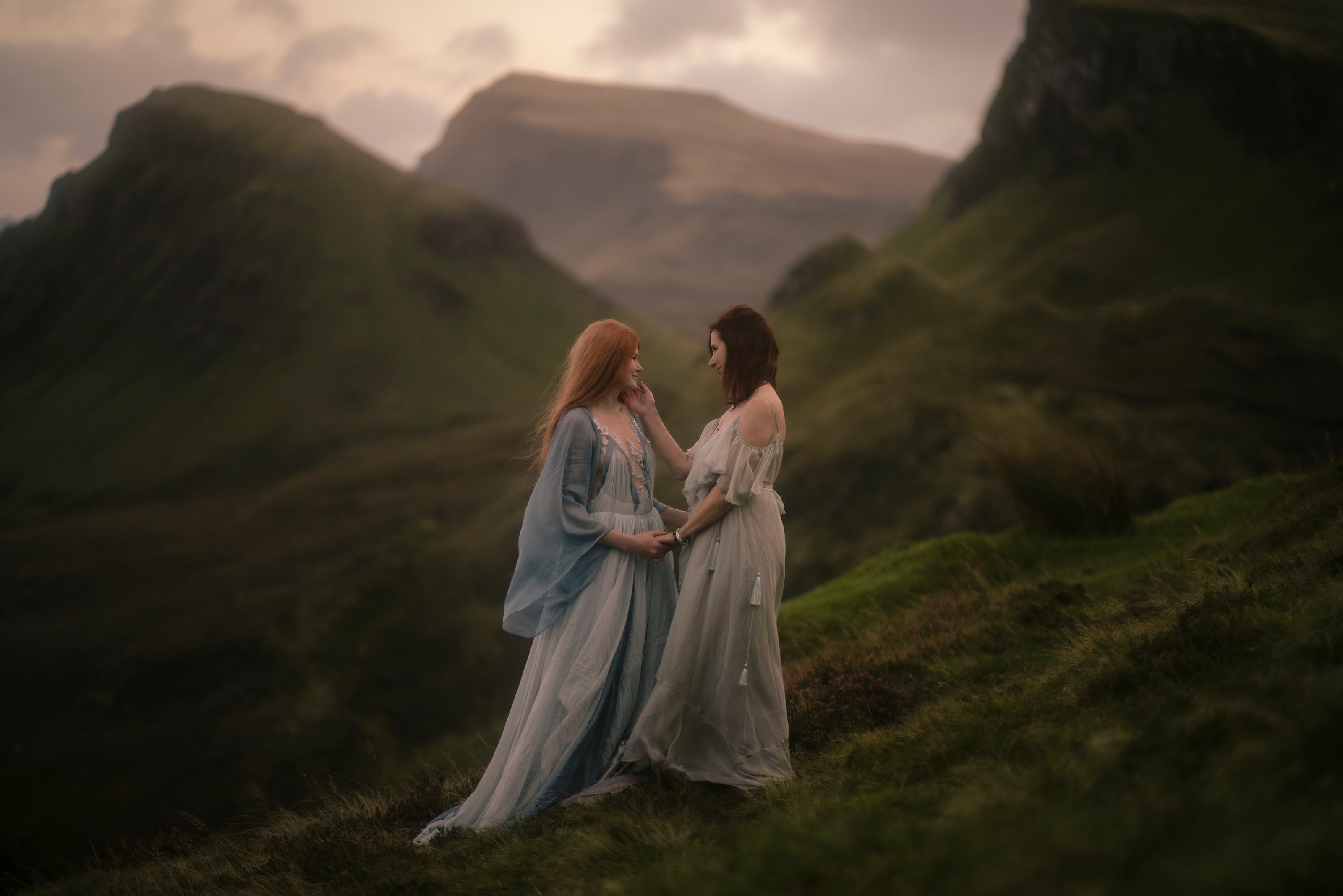 Mother and daughter in flowing dresses standing on a grassy hill with Scottish mountains in the background