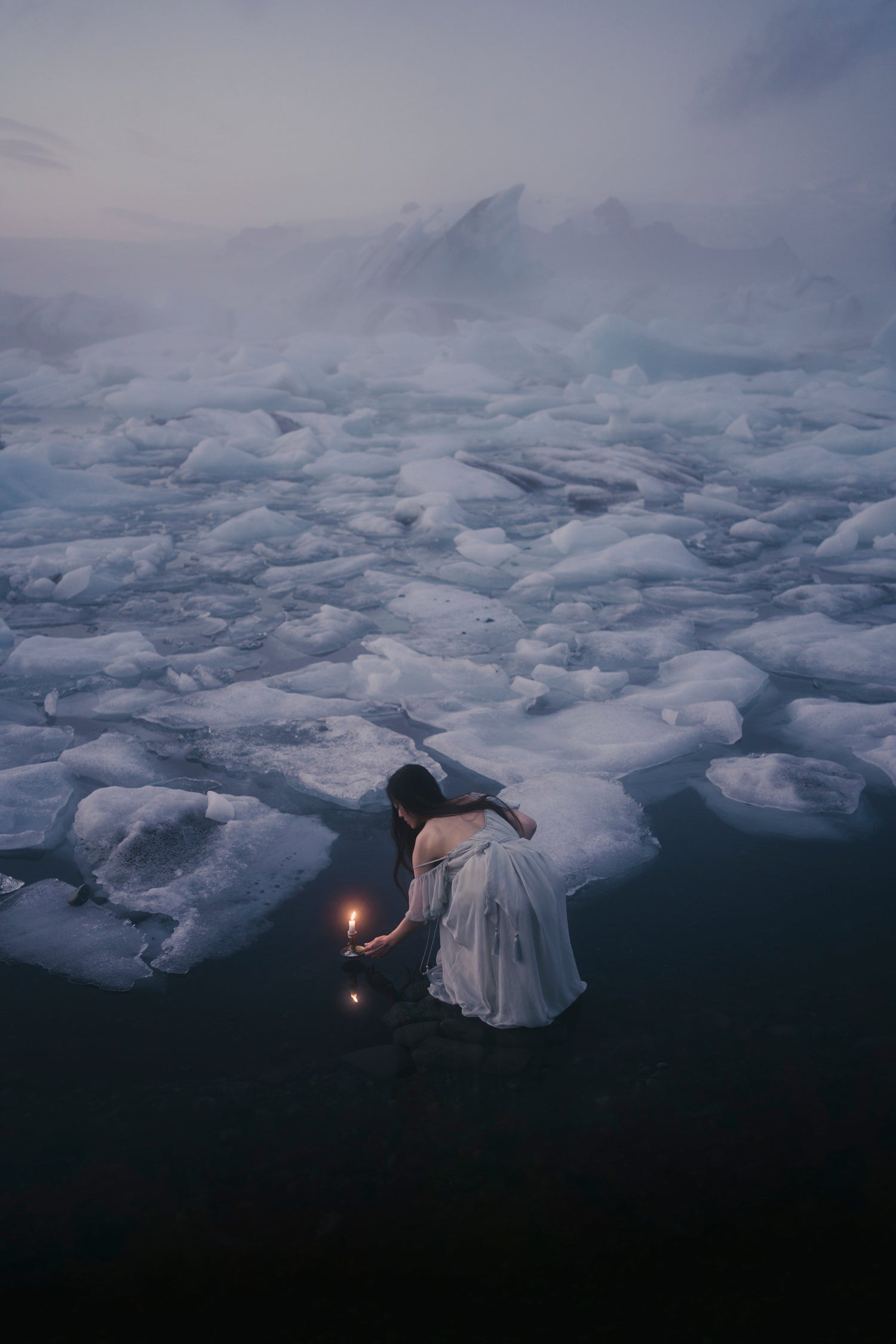 A woman in a long dress stands in dark water surrounded by floating ice, holding a small glowing candle. Mountains and mist fill the background in a moody, icy glacier lagoon.