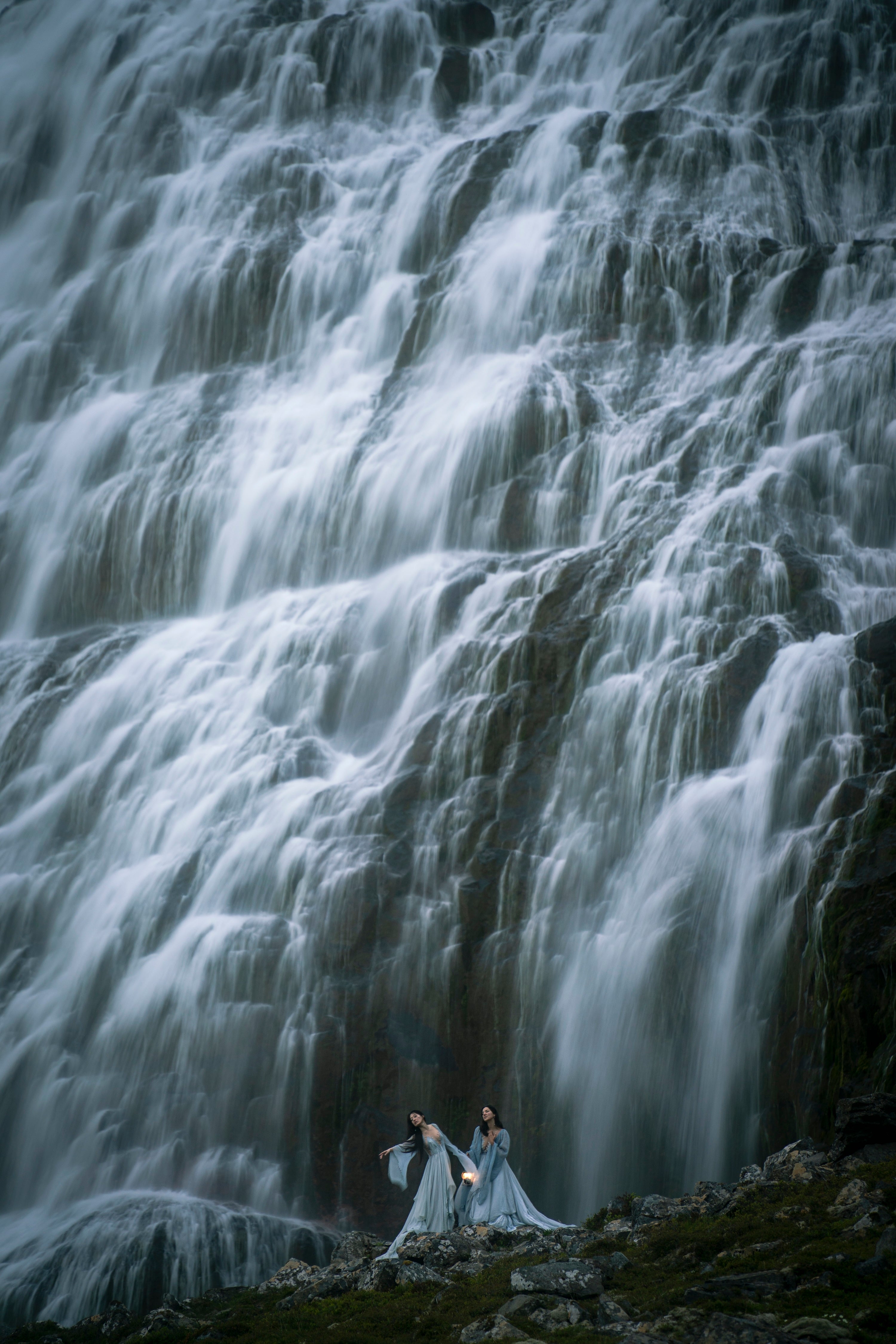 Two females in long silk dresses stand in front of a large flowing waterfall named Dynjandi