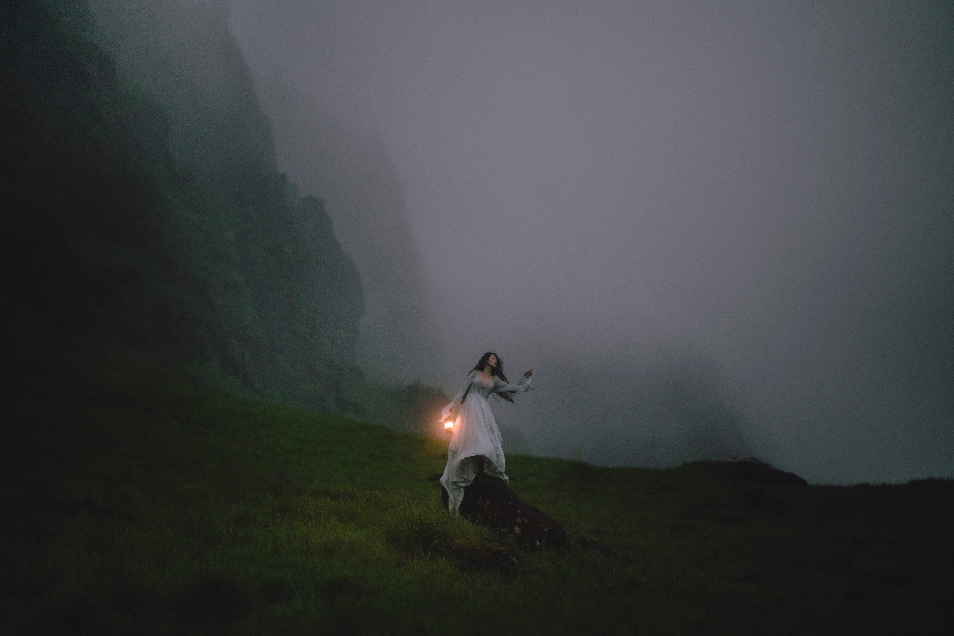 A woman in a long dress stands on a grassy hill holding a glowing lantern in a misty mountain landscape.