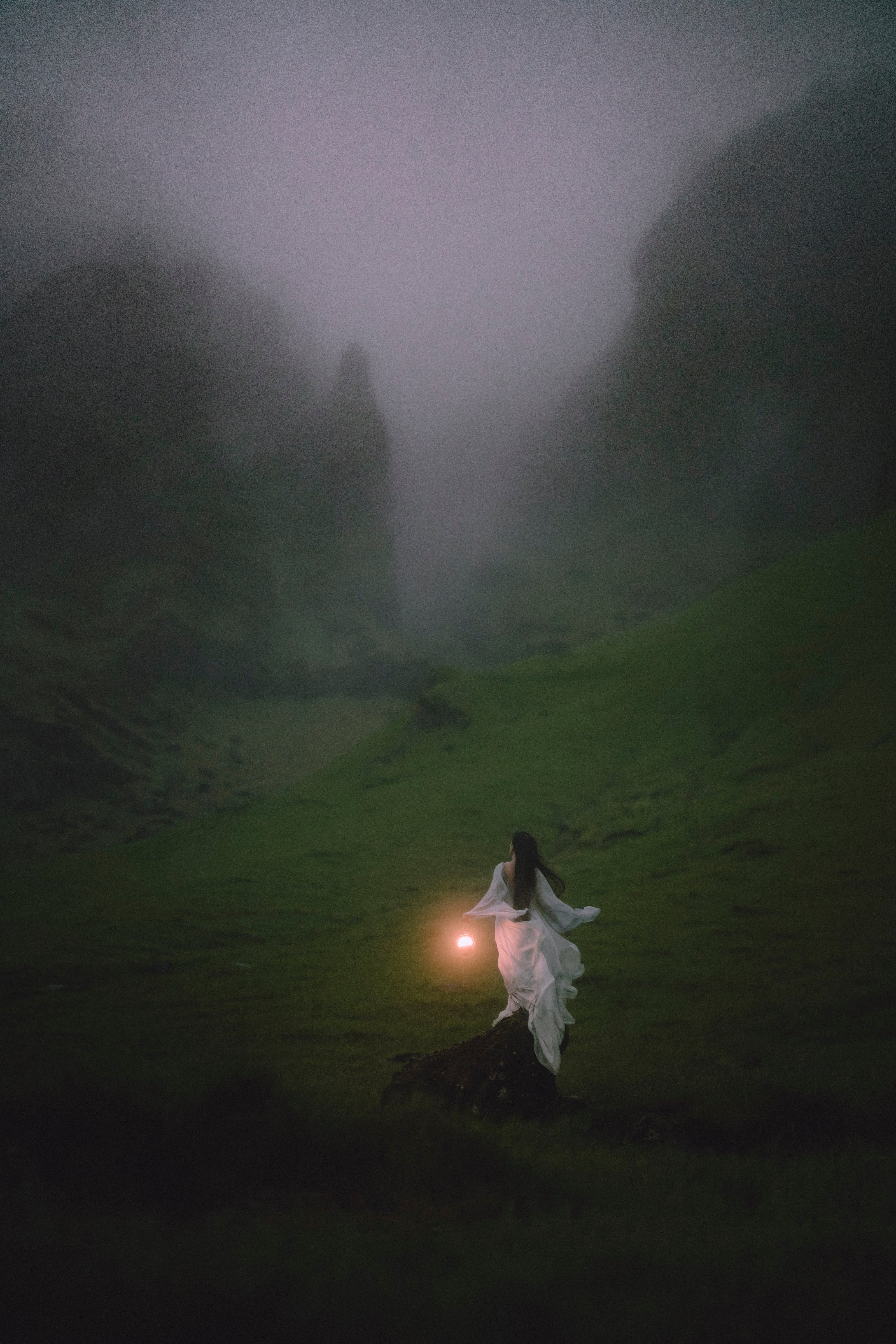 A person in a flowing MyWony dress stands on a dark hillside holding a glowing lantern in a misty green valley with tall shadowy Icelandic mountains in the foggy background.