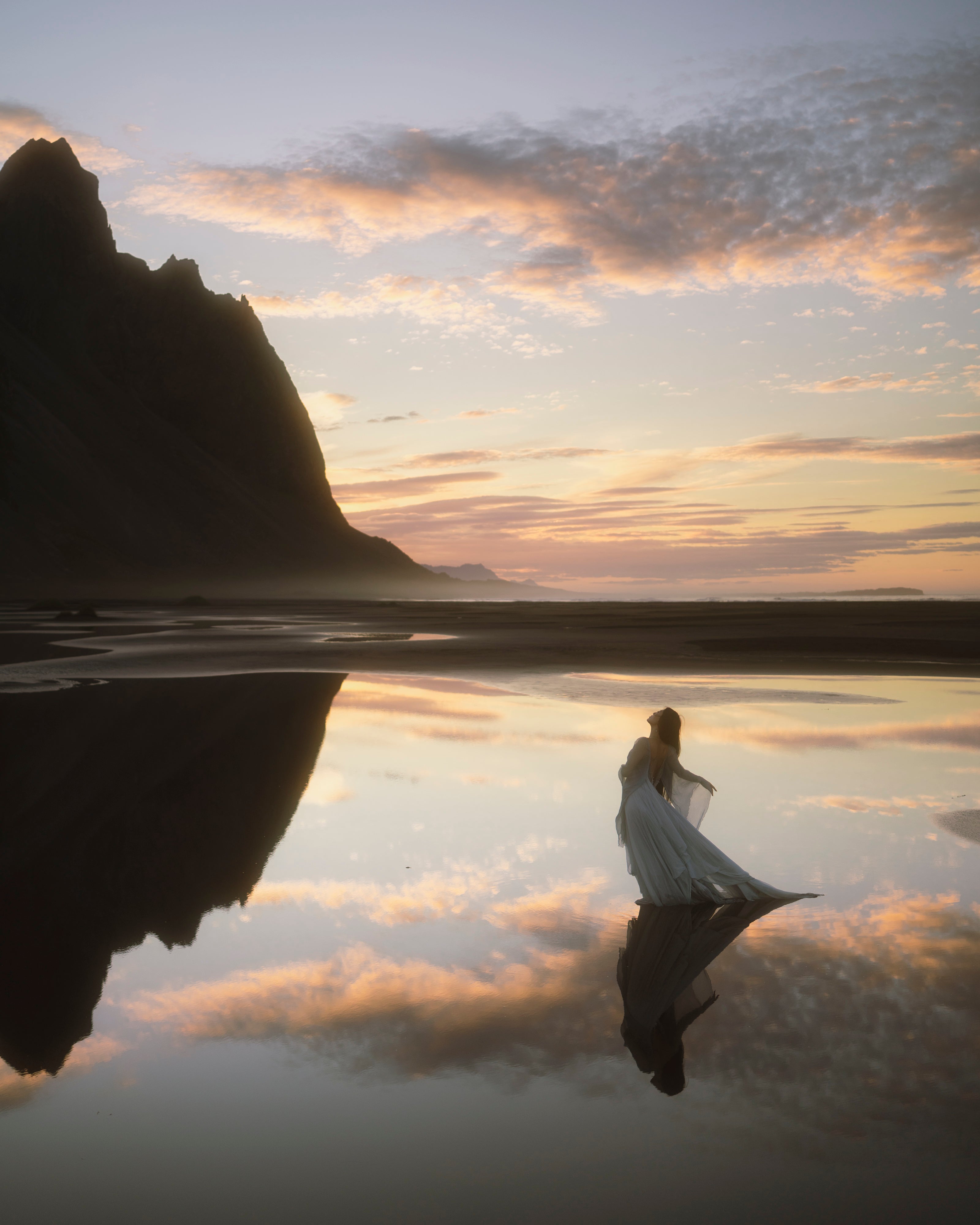 A woman in a flowing dress stands in shallow water at sunrise at Stokksnes, with a large dark mountain silhouette reflected in the calm surface. The sky is filled with soft orange, pink, and gray clouds.